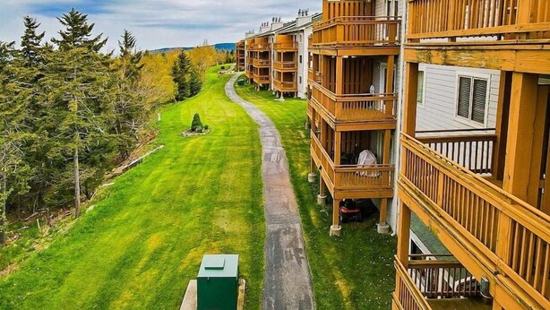 A row of wooden multi-story buildings with balconies, a paved pathway, and a grassy area with trees.