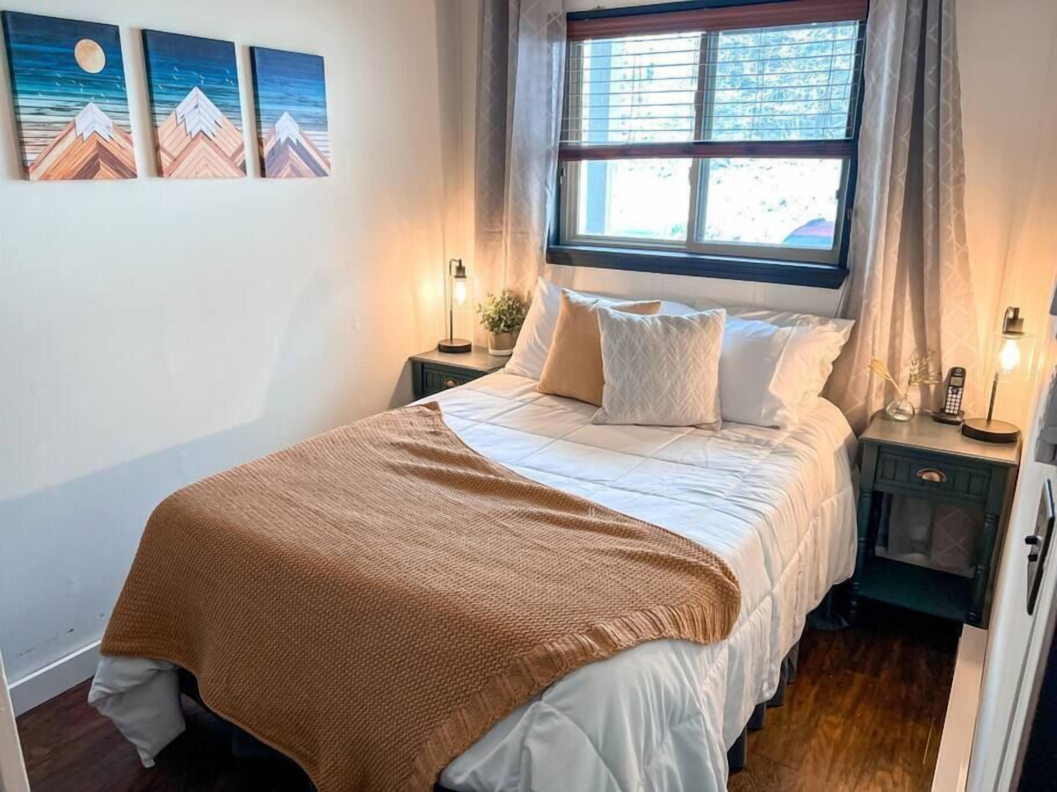 A neatly made bed with a brown blanket, a nightstand with a lamp, and a window with curtains.