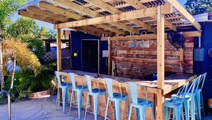 Un espace bar extérieur doté d’une pergola en bois, de chaises en métal bleu et d’un comptoir en bois.