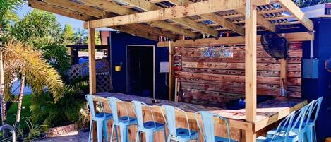 An outdoor bar area with a wooden pergola, blue metal chairs, and a wooden counter.