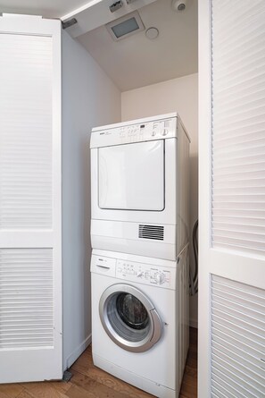 Une machine à laver et un sèche-linge superposés dans une buanderie, avec un plancher en bois et des murs blancs.