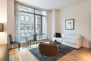 A modern living room with a white sofa, a round coffee table, and a dining table with chairs. Large windows offer a view of the city.