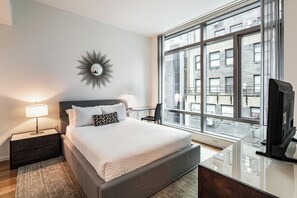 Habitación de hotel moderna con una cama grande, mesitas de noche con lámparas, un televisor y vistas de la ciudad a través de amplios ventanales.