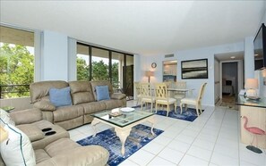 Relax in this stylish living room with a sofa, dining area, and kitchen, offering a view of lush greenery.