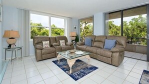 Sala de estar con dos sofás, una mesa de centro de vidrio y vistas a zonas verdes a través de grandes ventanales.
