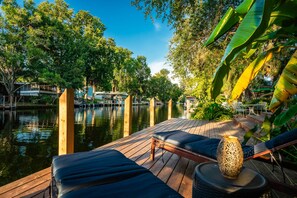 A wooden deck with lounge chairs and a decorative vase, overlooking a calm water body with trees and buildings in the background. - Waterfront Front Row Seats to Nature’s Best Show (Spring Hill)