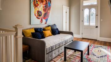 Un salon avec un canapé gris, des coussins colorés, une table basse noire, un tapis à motifs et un grand tableau représentant un chien accroché au mur.
