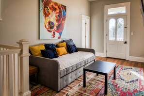 A living room with a gray sofa, colorful cushions, a black coffee table, a patterned rug, and a large painting of a dog on the wall.