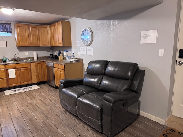 A living room with a leather sofa, wooden cabinets, a kitchen area, and a clock on the wall. - 1 BRM Basement Apartment/ 420 friendly outside (Lakewood)
