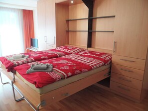 A double bed with a red and white checkered bedspread, a wooden wardrobe with multiple drawers, and a flat-screen TV on a small table.