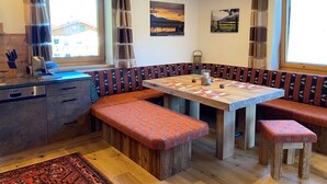 Ein gemütlicher Essbereich mit Holztisch und Bänken, eine Küche mit Spüle und Geschirr sowie ein Blick ins Freie durch große Fenster.