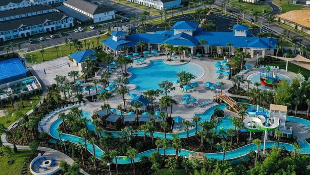 Luchtbeeld van een resort met meerdere zwembaden, palmbomen en een groot gebouw met een blauw dak.