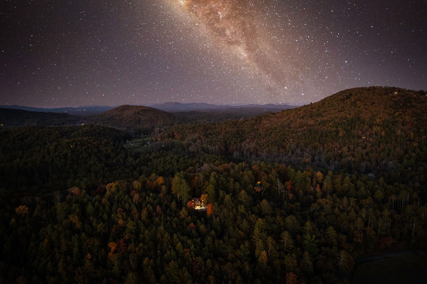 Una vista notturna di una zona boscosa con una piccola casa illuminata, circondata da colline e un cielo stellato.
