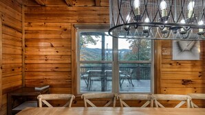 Interno di una baita in legno con tavolo, sedie, un lampadario e un'ampia finestra che si affaccia su una terrazza con mobili da esterno.