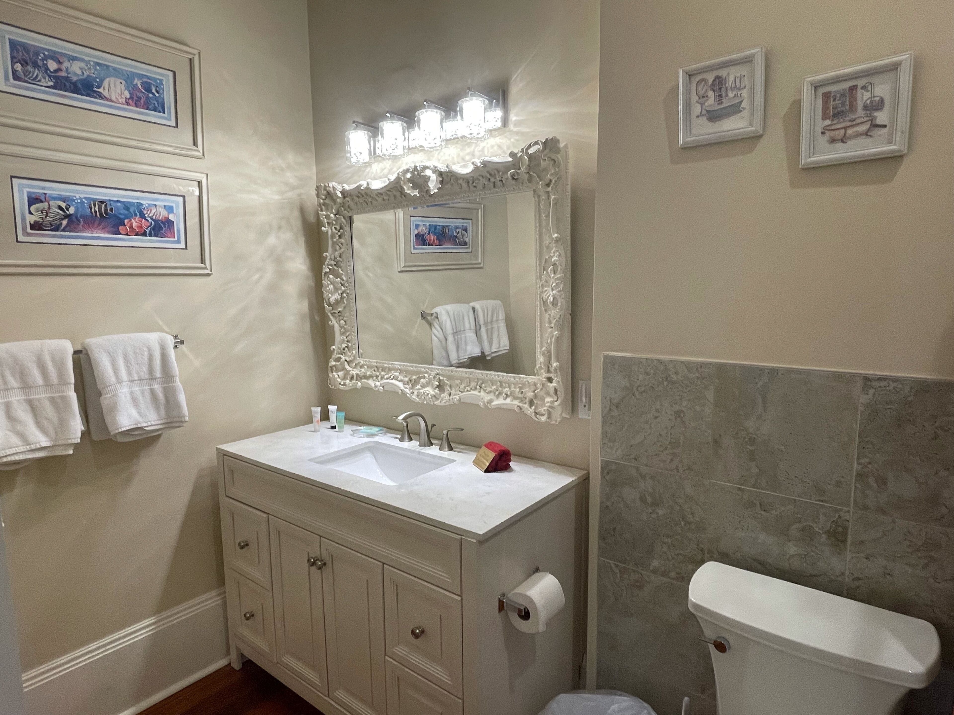 A bathroom with a white vanity, a large ornate mirror, and a toilet. There are towels hanging on the wall and framed pictures above the mirror.