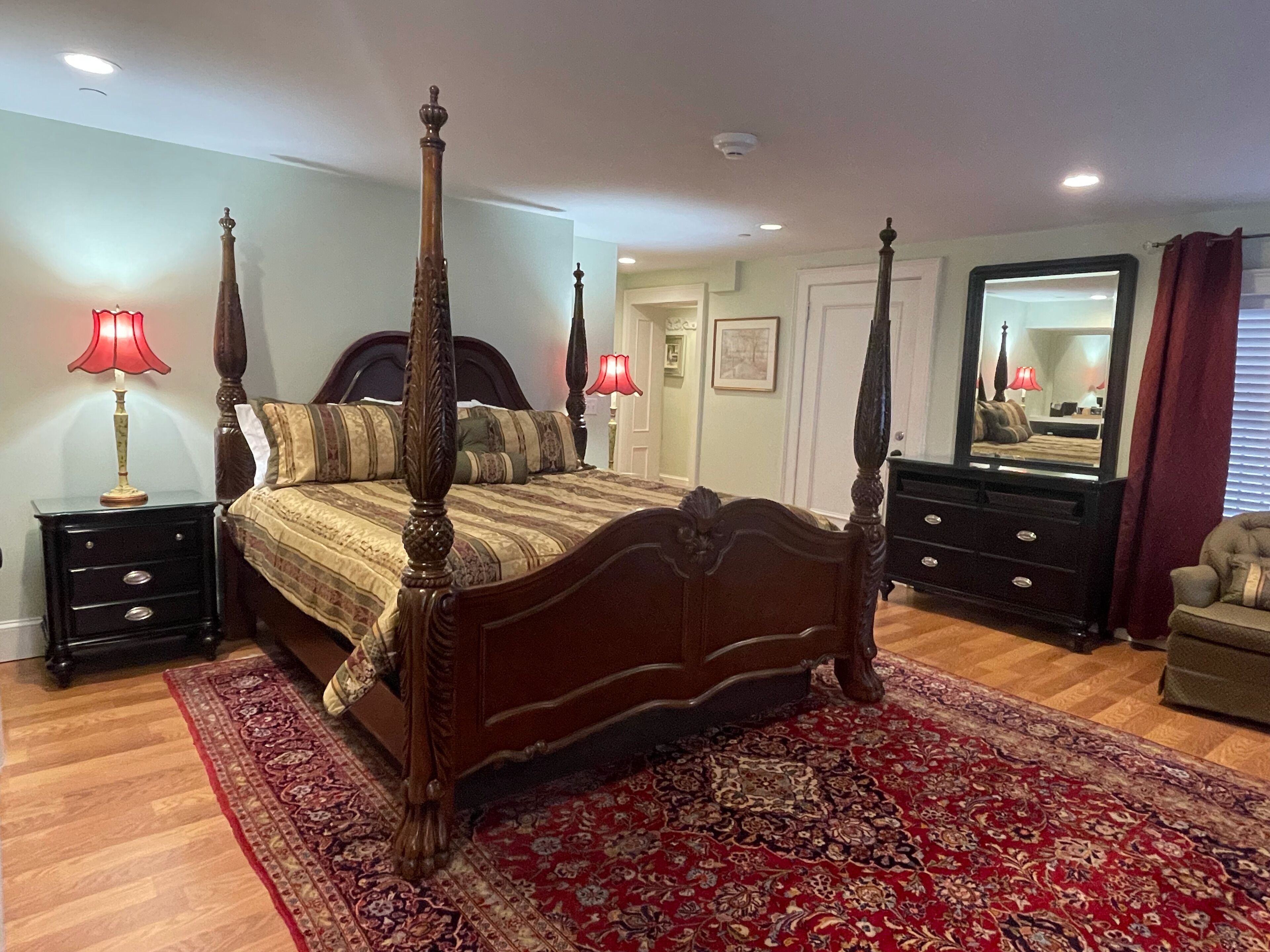 A four-poster bed with a red and gold patterned bedspread, flanked by two nightstands with lamps, a dresser, and a mirror.