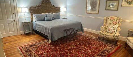 A bedroom with a large bed, a floral armchair, a red patterned rug, and framed pictures on the wall.