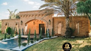 A brick building with a central arched entrance, surrounded by palm trees and small evergreen shrubs. - 🛡Medieval Castle Hôtel ™ (McAllen)