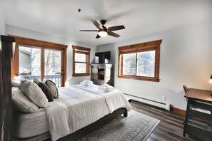 A bedroom with a bed, a ceiling fan, a fireplace, and large windows.