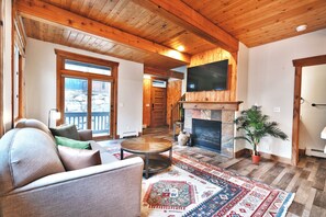 A spacious living room with a fireplace, a television mounted above it, and a large window offering a view of the outdoors.