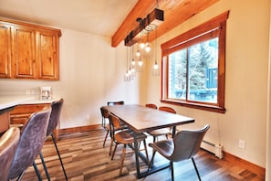 A dining area with a wooden table and chairs, a kitchen with wooden cabinets, and a window with a view of trees.