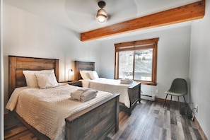 A bedroom with two beds, wooden headboards, a window with blinds, a chair, and a ceiling light.