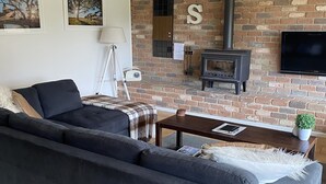 A living room with a brick wall, a sofa, a table, a TV, and a fireplace. - Farm stay with alpacas at your doorstep (Cromer)