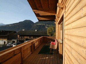 Un balcon en bois offrant une vue sur une chaîne de montagnes, un tapis vert et une chaise rouge.
