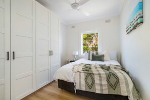 A bedroom with a bed, a window, a ceiling fan, and a painting on the wall. - Little Manly Getaway (Manly)