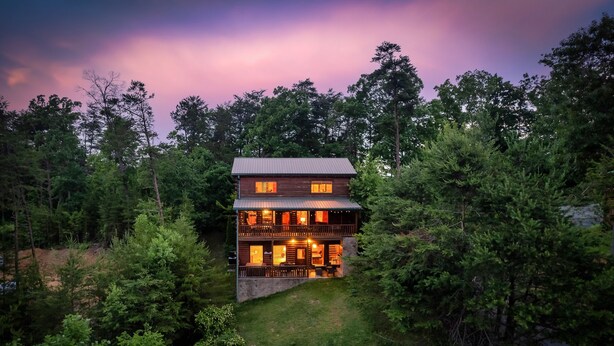 A two-story log cabin with a balcony, surrounded by dense forest. - Log Cabin with Stunning Views- Hot Tub, Sauna, BBQ, Patio Fireplace, Pets ok (Sevierville)