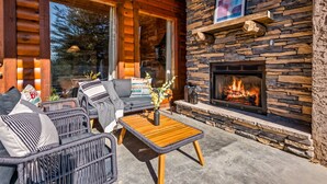 A cozy outdoor seating area with wicker furniture, a stone fireplace, and a large window overlooking a forested area. - Log Cabin with Stunning Views- Hot Tub, Sauna, BBQ, Patio Fireplace, Pets ok (Sevierville)