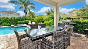 A covered patio with a glass-top table and wicker chairs, a barbecue grill, and a swimming pool.