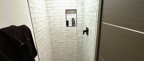 A modern bathroom with white brick walls, a glass shower enclosure, a black towel on the wall, and a wooden floor.
