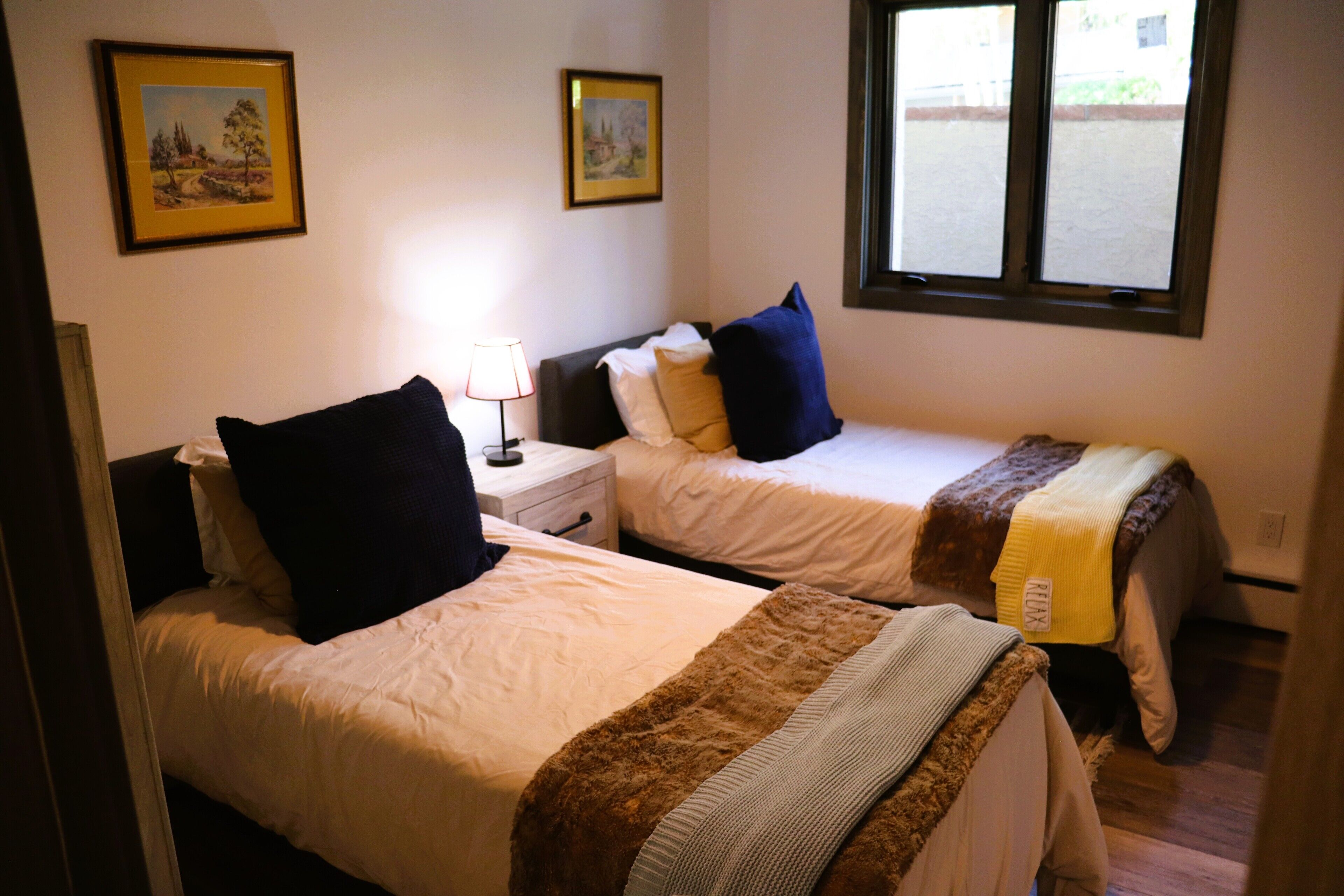 A room with two beds, a nightstand, a lamp, and framed pictures on the wall.