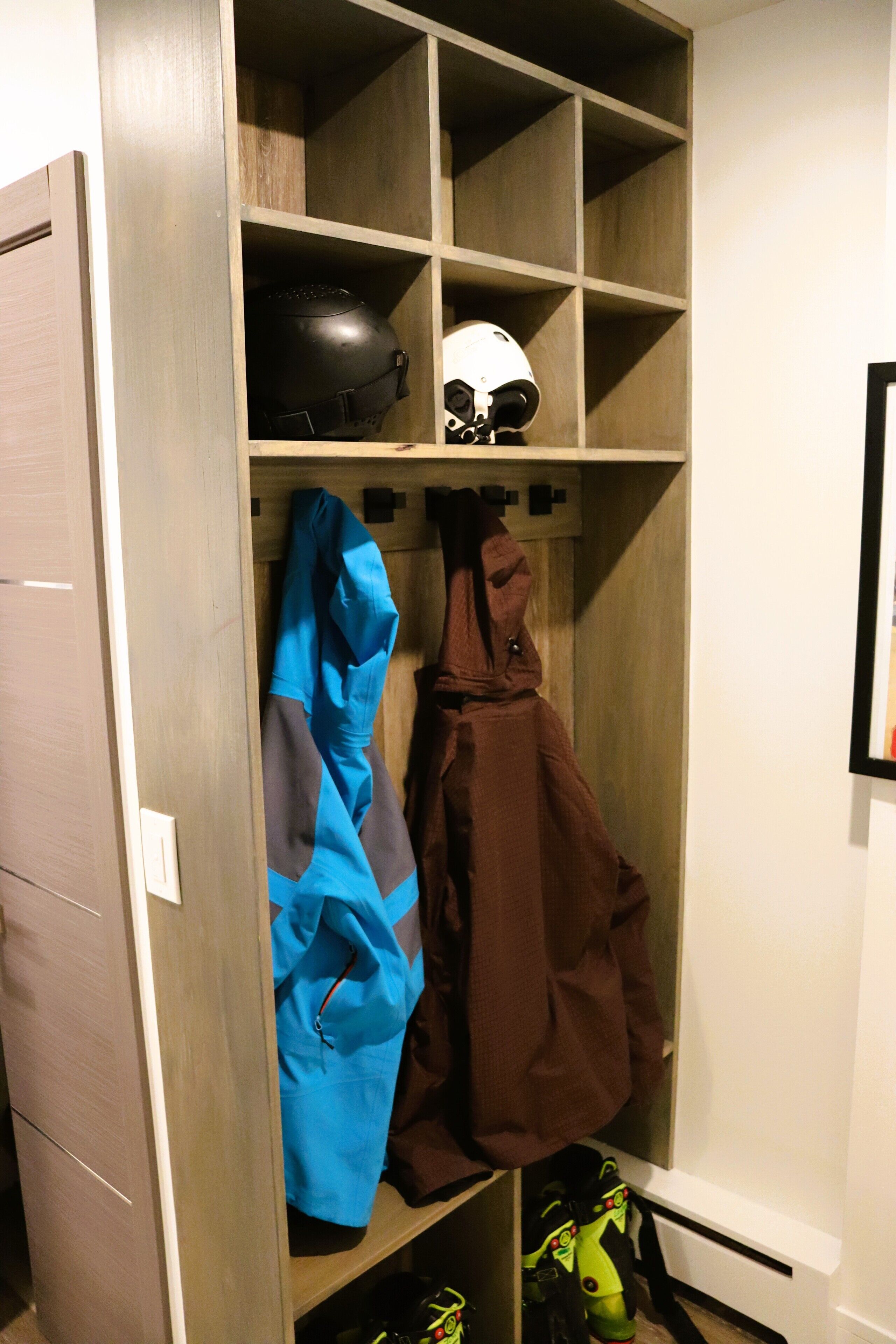 Un placard en bois, doté d’étagères et de crochets, contenant un casque, une veste bleue et une veste marron.