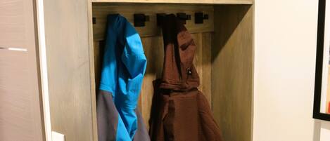Een houten kast met planken en haken, met een helm, een blauw jasje en een bruine jas.