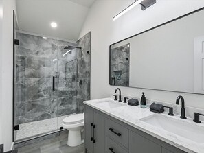 A modern bathroom with a glass-enclosed shower, a white toilet, and a marble countertop with black fixtures. - Newly Renovated Luxury 5BR – Family & Game Room (The Colony)