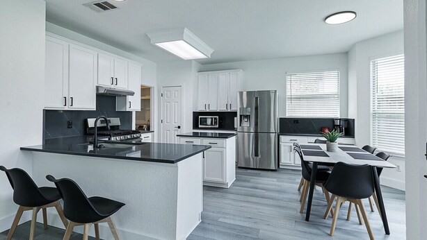 A modern kitchen with white cabinets, a black countertop island, stainless steel appliances, and a dining area with black chairs. - Newly Renovated Luxury 5BR – Family & Game Room (The Colony)