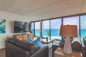 A modern living room with a large TV, comfortable seating, a coffee table, and a view of the ocean.