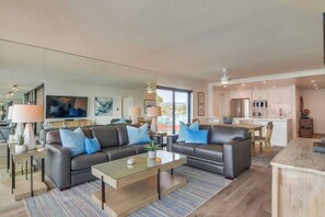A modern living room with a glass coffee table, two sofas with blue cushions, a flat-screen TV, and a large mirror.