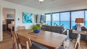 Een moderne woonkamer met een houten eettafel, comfortabele zithoek, een flatscreen-televisie en een groot raam met uitzicht op de oceaan.