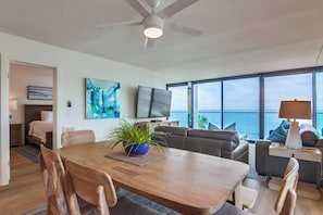 Een moderne woonkamer met een houten eettafel, comfortabele zithoek, een flatscreen-televisie en een groot raam met uitzicht op de oceaan.