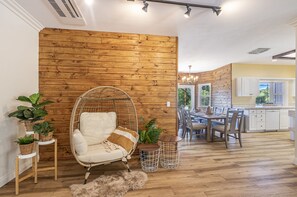 Relax in a cozy seating area with a wicker chair and plush rug, adjacent to a dining space and kitchen with a view of the outdoors.