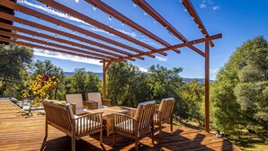 Een houten terras met een pergola, tuinmeubilair en uitzicht op bomen en heuvels.