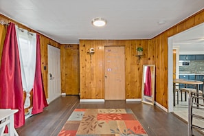 A hotel room with wooden walls, a wooden door, a red curtain, a patterned rug, and a small plant on the wall.