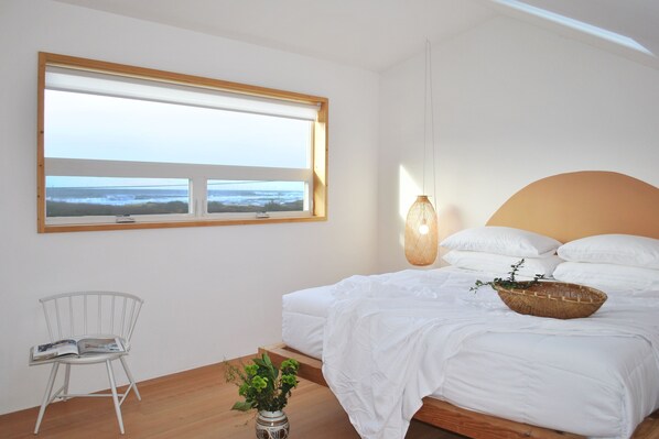 A bedroom with a large window, a bed with white bedding, a wicker basket on the bed, a white chair, and a floor lamp. - Ocean View, Perfect for Agate Hunting! Dog Friendly! Fireplace! (Florence)