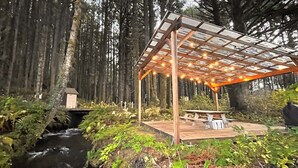 Una terraza de madera con un pérgola y luces colgantes, con vistas a un arroyo rodeado de bosque denso.