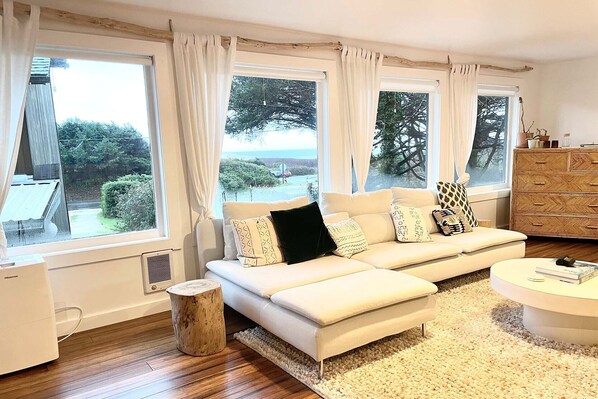 A modern living room with a sofa, a round coffee table, and a wooden stool. There are large windows with curtains, a view of greenery outside, and a wooden cabinet.