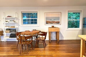 Uma área de refeições com mesa e cadeiras de madeira, uma estante branca com livros e uma cesta, e duas janelas com vista para o exterior.
