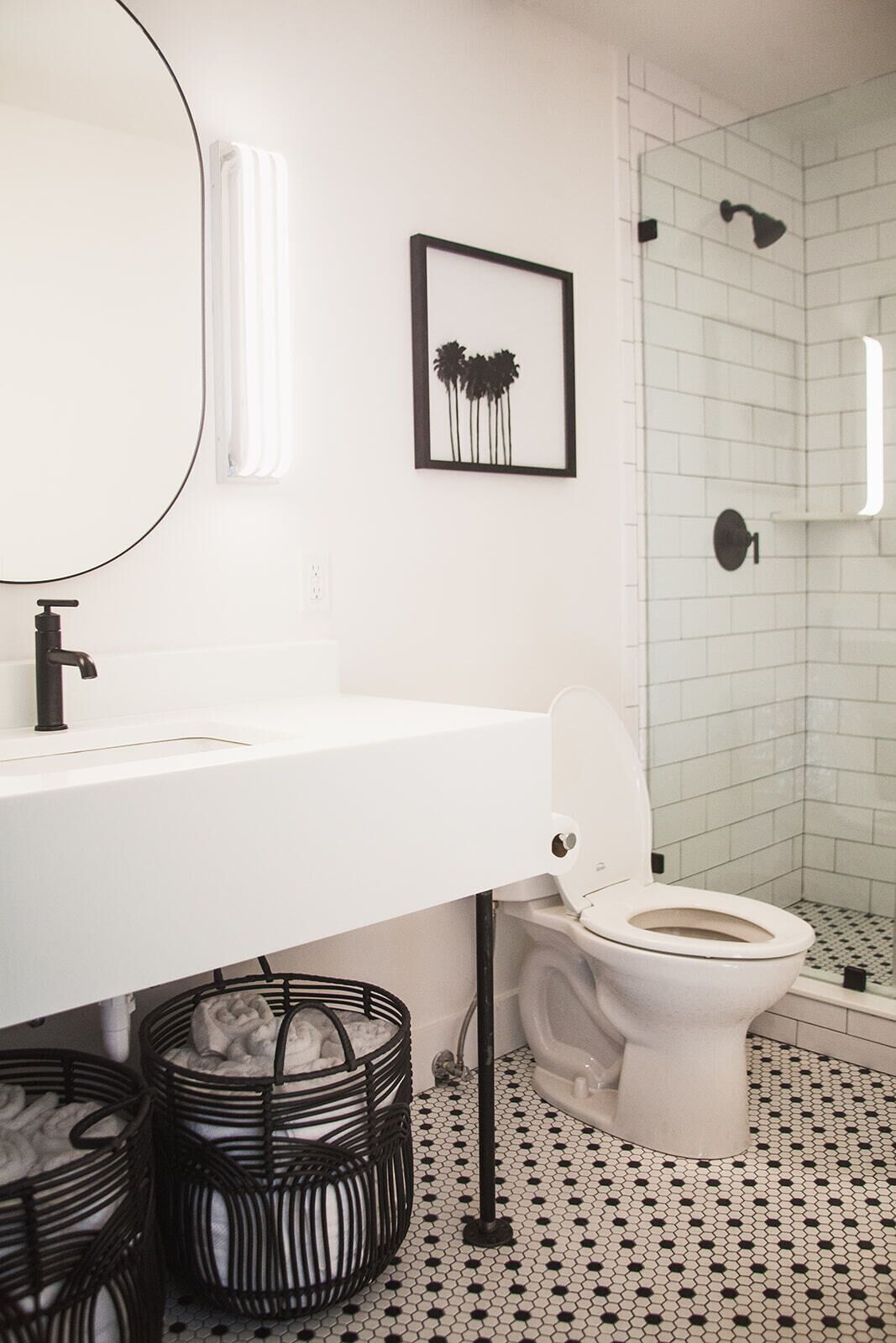 Une salle de bain avec un lavabo, des toilettes et une douche, le tout de couleur blanche. Un tableau encadré est accroché au mur et un panier noir se trouve sous le lavabo.